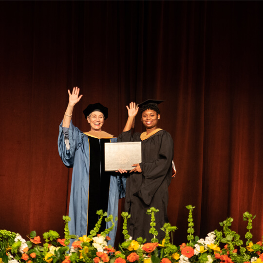 Dean Mills waves on stage while presenting graduate with diploma