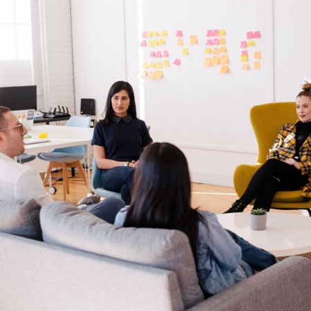 Two women sit on couches and talk near whiteboard with post-it notes.